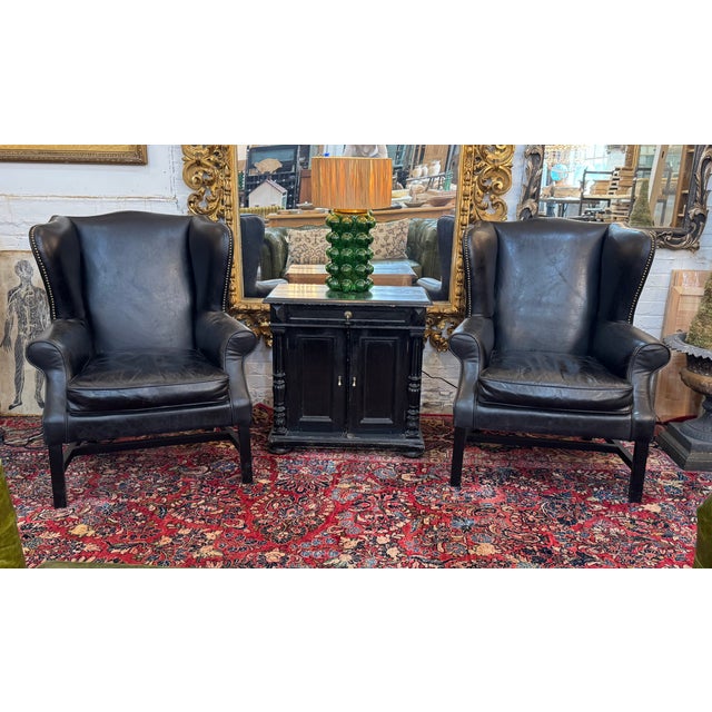 Stylish pair of vintage wingback chairs upholstered in black leather. Features classic high backs, gently rolled arms, and...