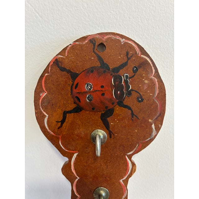 A delightful vintage folk art wall hook rack featuring a hand-painted red ladybug on a warm-toned wooden plaque with...