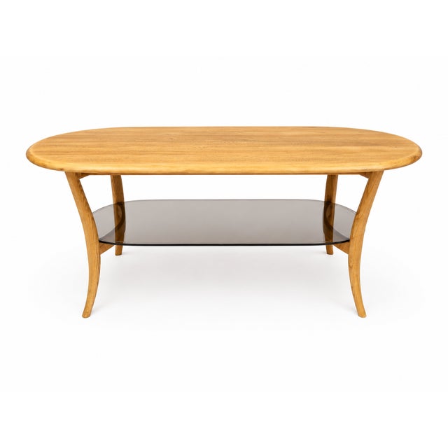 A beautiful Ercol model 1063 oval coffee table, featuring a blonde wood finish, elegant curved legs and a smoked glass...