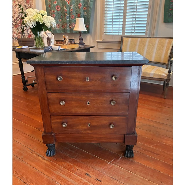 A French Empire three-drawer commode, circa 1810–1830, with tailored rectangular form and paw-foot base. Crafted from...