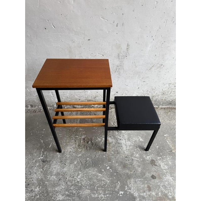 A stylish mid-century modern side table featuring a teak top and an integrated wooden slat magazine rack. The clean black...