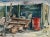 Rendered in deep shades of green and brown, the pile of raw materials and trash beside a metal dumpster appears rustic and...