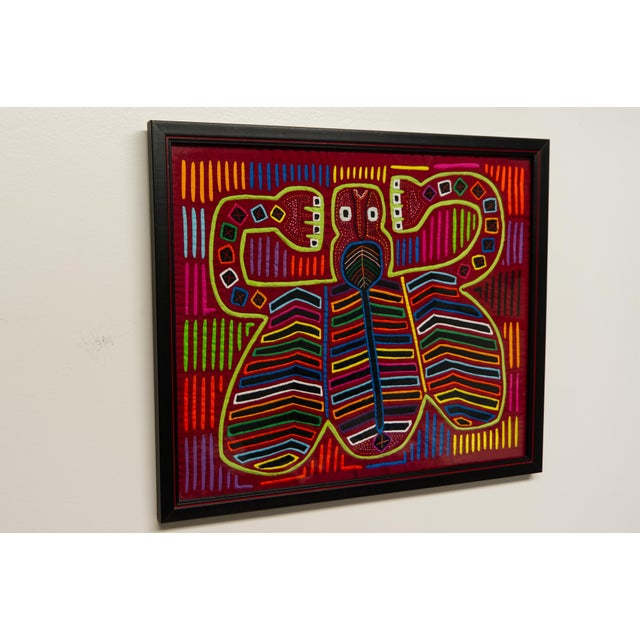 A framed textile mola art hand crafted by the Kuna women of San Blas, Panama. Black wood frame with red line detail....
