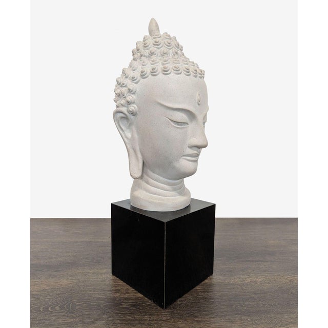 This Buddha head sculpture features a calm and peaceful expression, crafted in a light-toned material. It is mounted on a...