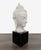 This Buddha head sculpture features a calm and peaceful expression, crafted in a light-toned material. It is mounted on a...