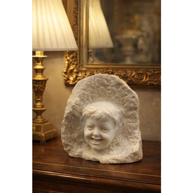 A sculpture depicting a child's face emerging from a block of marble. The contrast between the smooth face and the rough...