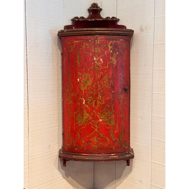 A hanging corner cupboard with a bowed front, painted overall on a red ground with gilt scrollwork cartouches enclosing...