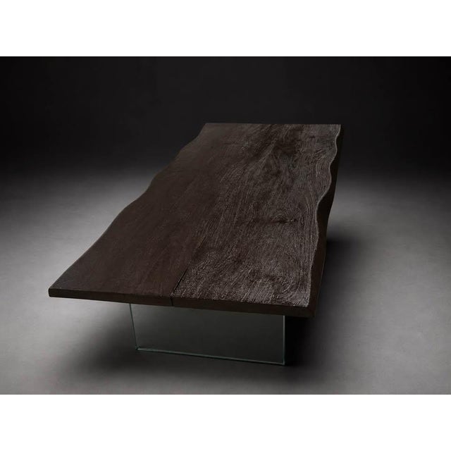 A modern live edge-style coffee table featuring a dark textured wood-look top and clear glass panel legs. The combination...