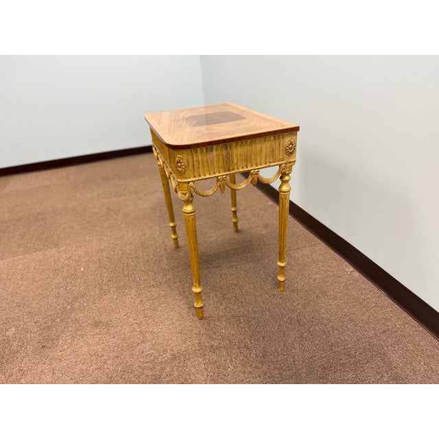 Elegant Louis XVI–style giltwood side table featuring fluted legs, decorative swag detailing, and a warm wood veneer top....