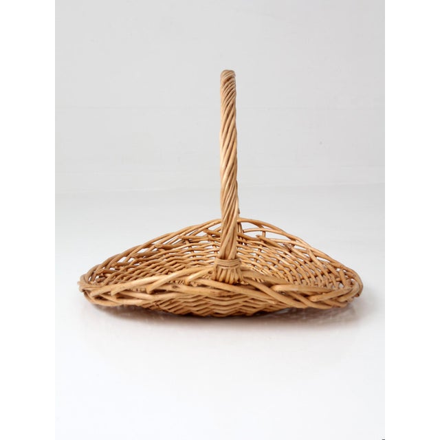 Large vintage wicker gathering basket with a wide, open weave and a tall arched handle that creates a strong, sculptural...