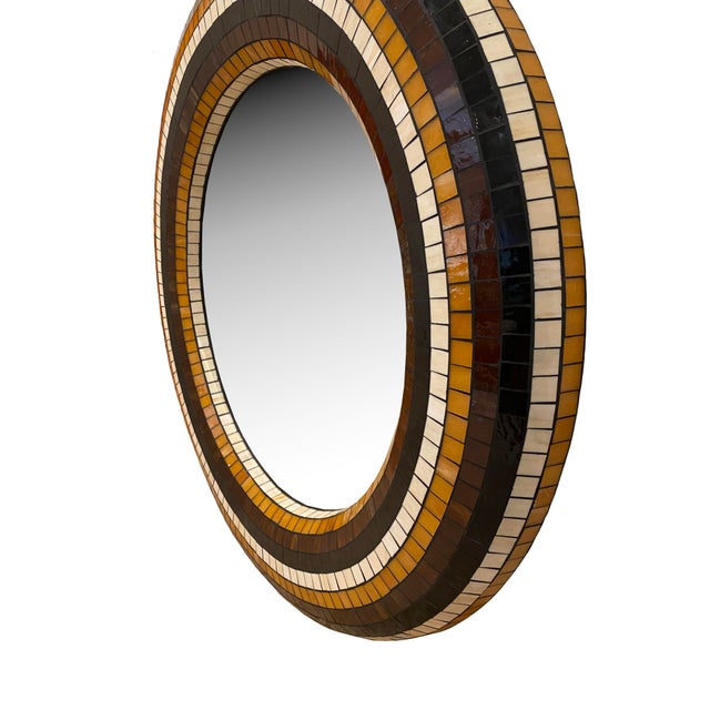 A large round mirror framed in glass mosaic tiles, blending classic Italian hues of yellow, black, and burgundy. It has...