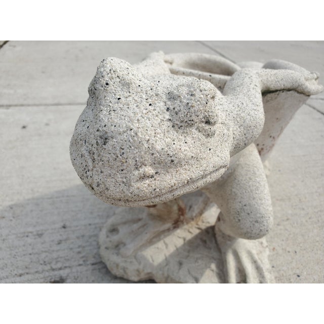 This item is a decorative concrete garden statue featuring a frog pulling a snail or nautilus shell, which functions as a...