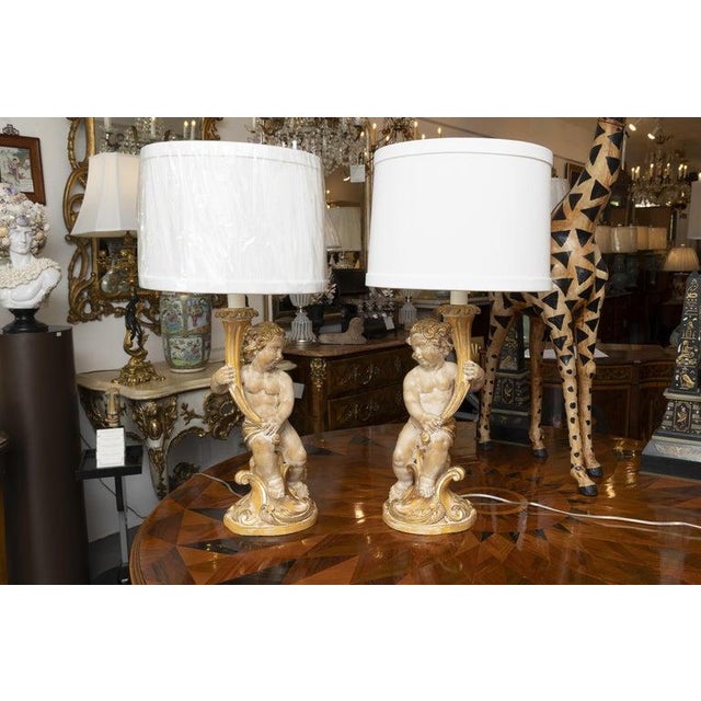 This is a charming pair of Italian giltwood candlestick lamps modeled as kneeling cherubs supporting foliate...