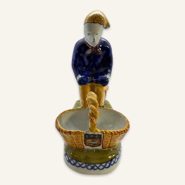 This Quimper salt cellar/condiment dish represents a man squatting over a basket to perform a very private function....