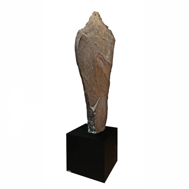 A tall Stone sculpture hand-carved in Indonesia, mounted on a black metal cube stand. The slate is narrow on the bottom,...