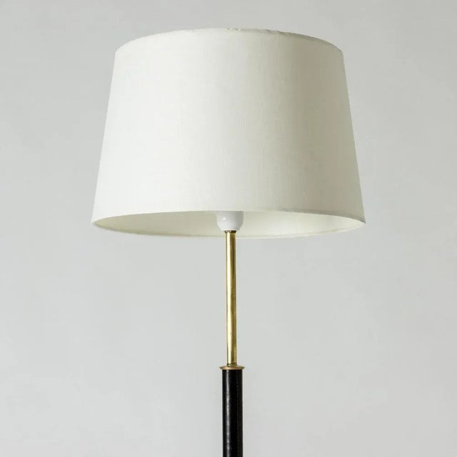 Elegant midcentury floor lamp, made from brass with a leather dressed stem. Black lacquered base. European Plug (up to...