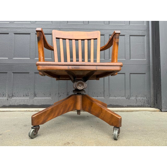 Classic Early 20th Century wooden banker’s chair featuring a curved slatted back, sculpted seat, and solid wood arms. This...