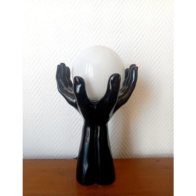 Vintage lamp, circa 1970, depicting two black glazed ceramic hands holding a luminous white opaline glass globe. Clean and...