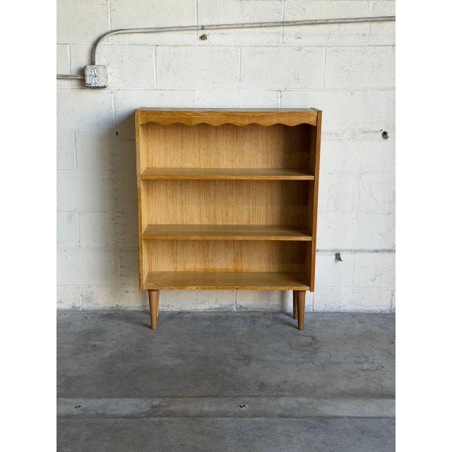This oak bookcase has a compact upright profile with three open display levels. A scalloped trim runs beneath the raised...