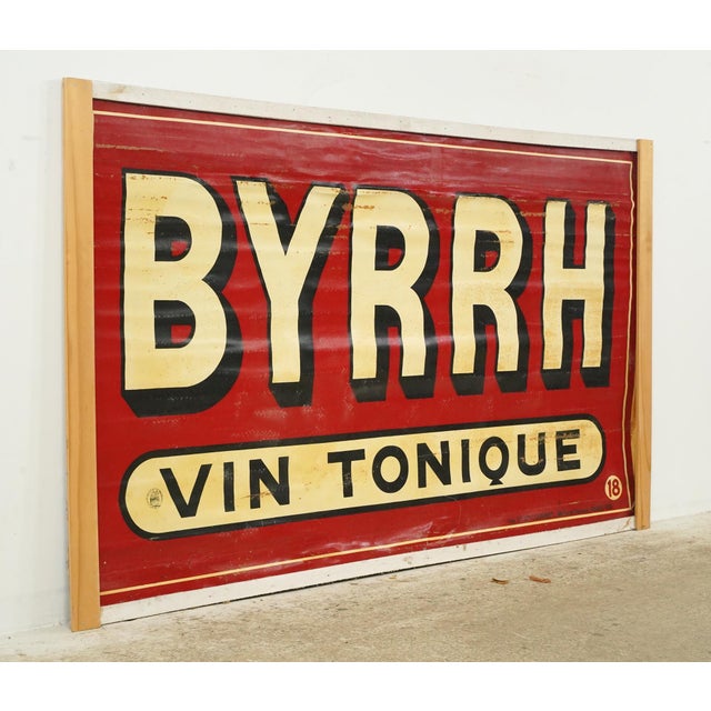 The sign is painted on a red background with off white colored letters that reads "BYRRH VIN TONIQUE" which translates to...