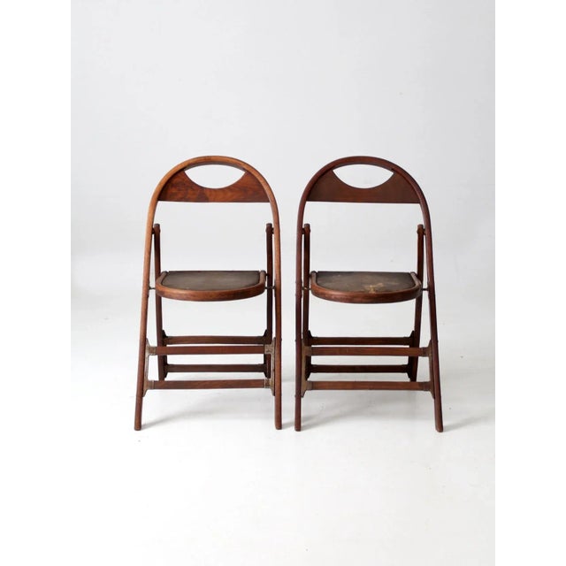 A distinctive pair of antique wooden folding chairs, dating to the early 20th century. The chairs feature gracefully...