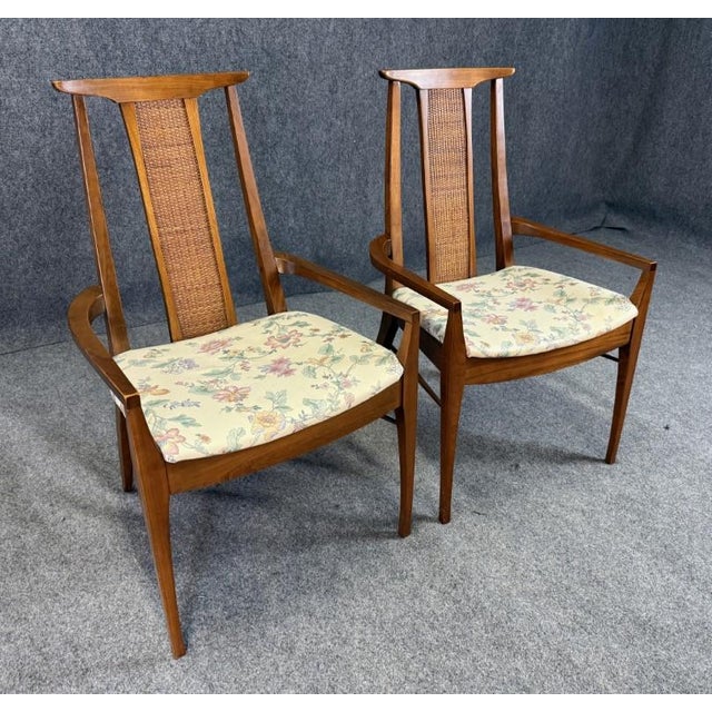 This pair of mid-century modern walnut arm chairs features gently curved frames with warm wood tones and tall cane back...