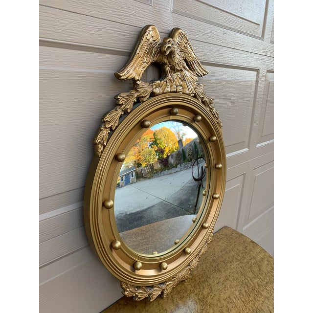 A Federal-style giltwood convex mirror crowned with a carved eagle, its wings outstretched above a laurel motif—an...