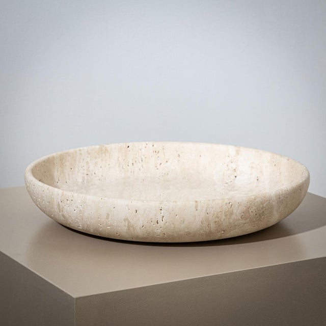 A large, heavy stone bowl crafted from beige travertine, featuring a minimalist design. Its gently sloping rim and flat...
