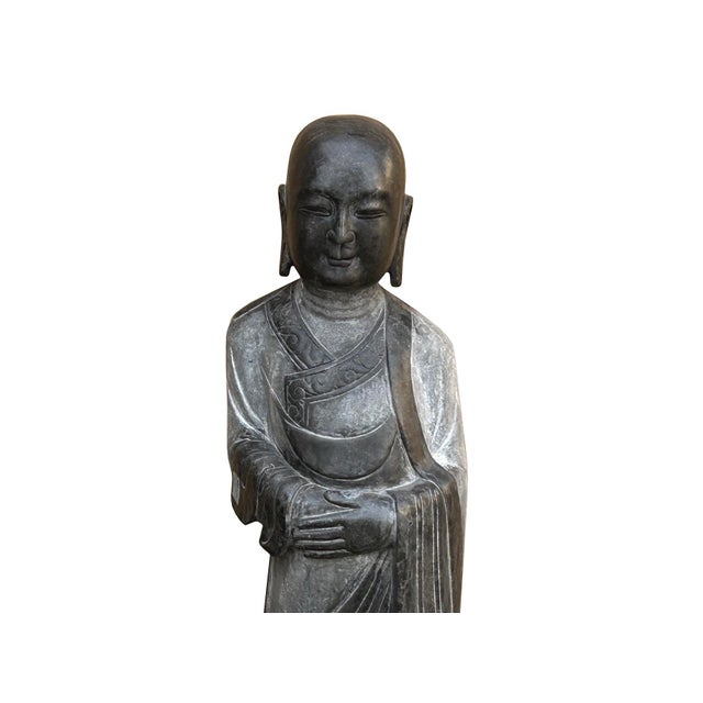 This striking hand-carved dark gray stone statue depicts a Buddhist monk standing in serene contemplation atop a double-...