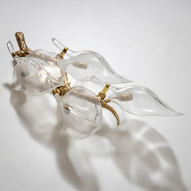 Wall lamp in the shape of a stylized branch, crafted in brass and adorned with four large clear-glass leaves featuring...
