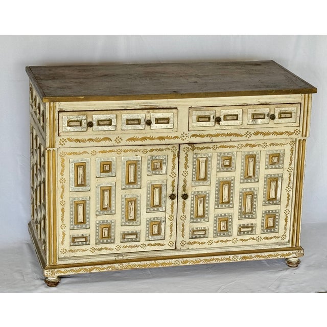 A two-door, two-drawer cabinet. The surface painted in an off-white with gold decorations. Adorned with raised panels, the...