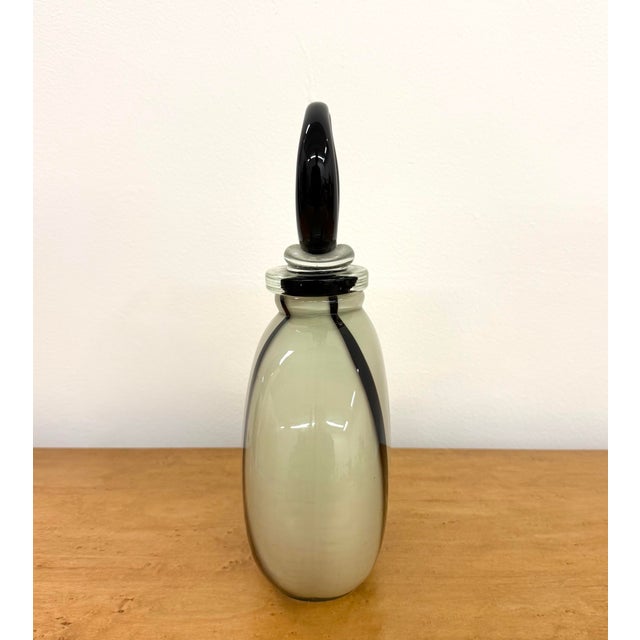 A vintage hand-blown art glass decanter featuring a rounded ivory body accented by deep black vertical bands and applied...