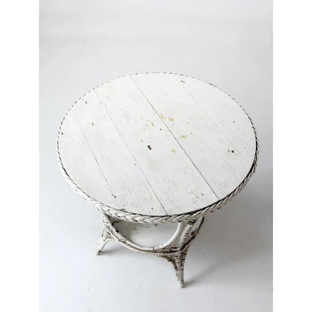 An antique wicker accent table with a round painted wood top, woven wicker base and lower stretcher shelf. The timeworn...