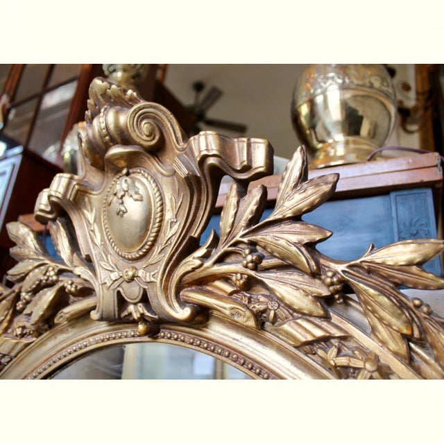 A boldly crested antique mirror with a ribboned cartouche and laurel branches, the gilded oval frame is composed of a...