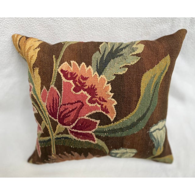 An exquisite antique-style pillow upholstered in richly woven 19th-century tapestry, set against a deep warm brown ground...