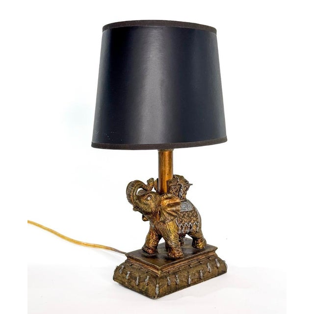 A gold and silver highlighted bronze toned petit resin figural table lamp featuring an elephant with his trunk showing in...