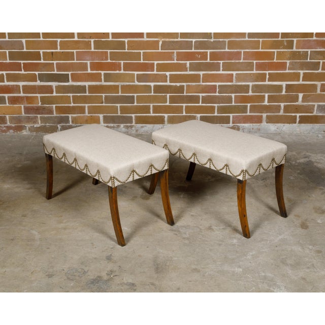 A pair of English Regency period mahogany rectangular stools from the 19th century with saber legs and custom upholstery....
