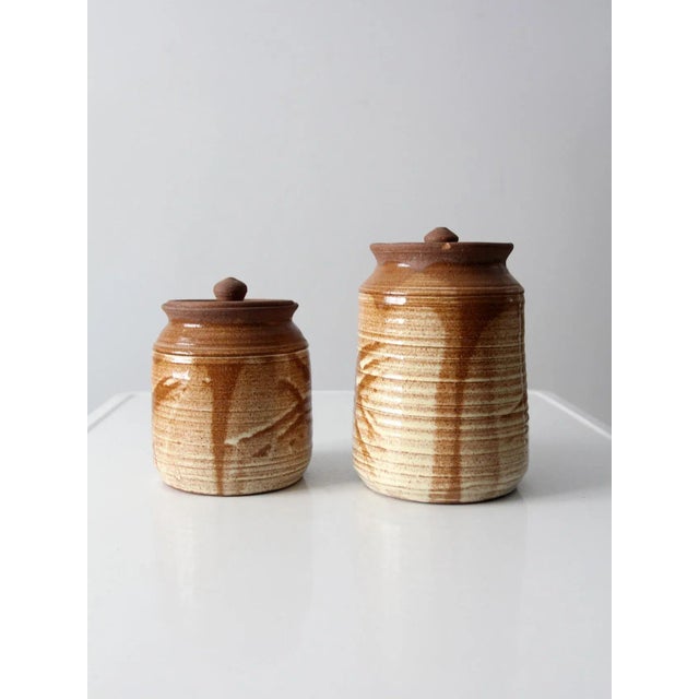 This is a pair of vintage studio pottery jars. Signed by the artist, the hand-crafted jars feature a warm speckled brown...