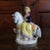 A couple riding finely modeled prancing white horses, each figure in elegant attire, the gentleman in a military jacket....