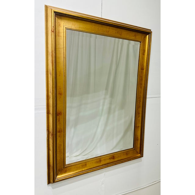 A striking contemporary wall mirror featuring a wide carved giltwood-style frame with warm antiqued gold finish and clean...