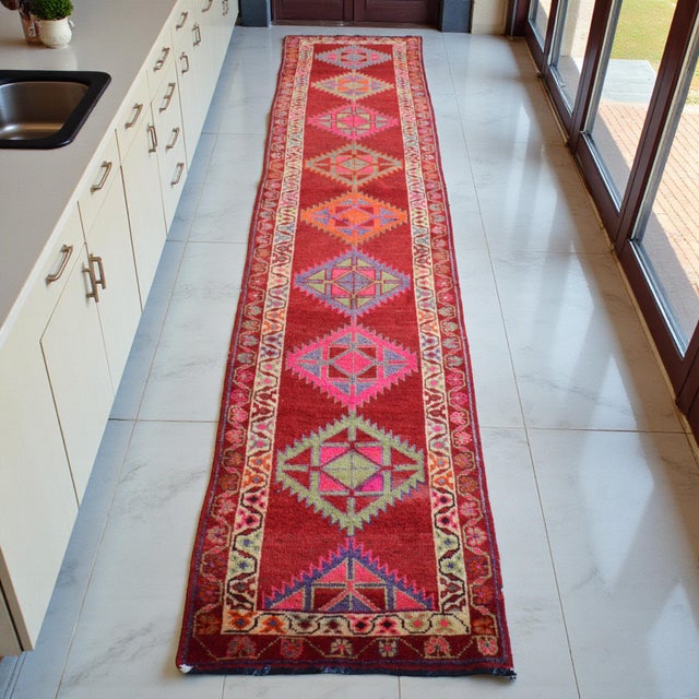 This vibrant 1970s Oushak runner features a series of striking geometric medallions set against a rich, saturated red...
