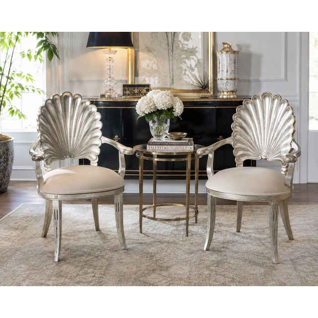 A glamorous pair of Hollywood Regency shell back armchairs featuring sculpted scallop-form backs, an antiqued silver leaf...