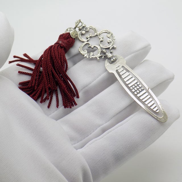 This vintage silver reading accessory depicts an elegant bookmark finished with a decorative red tassel, crafted as a...
