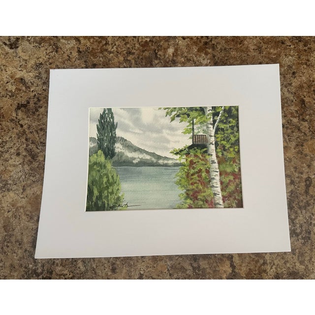 This serene landscape watercolor captures a peaceful lakeside scene with a birch tree, leafy shoreline, and distant...