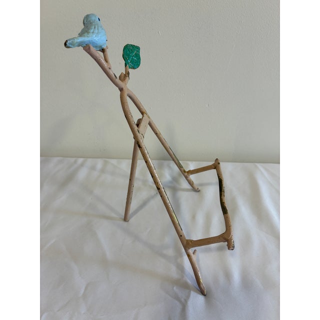 A charming vintage cast iron easel display stand featuring a hand-painted blue bird perched on a decorative branch with a...