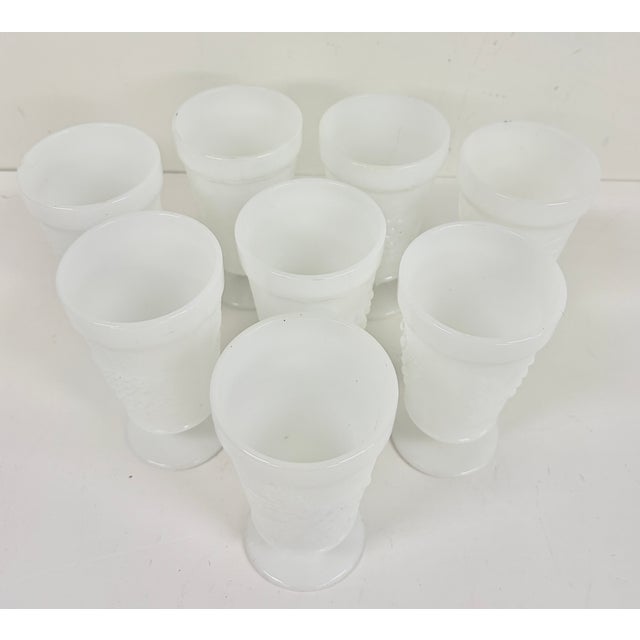 Set of eight vintage white milk glass goblets featuring a raised textured pattern, flared rims, and pedestal bases. The...