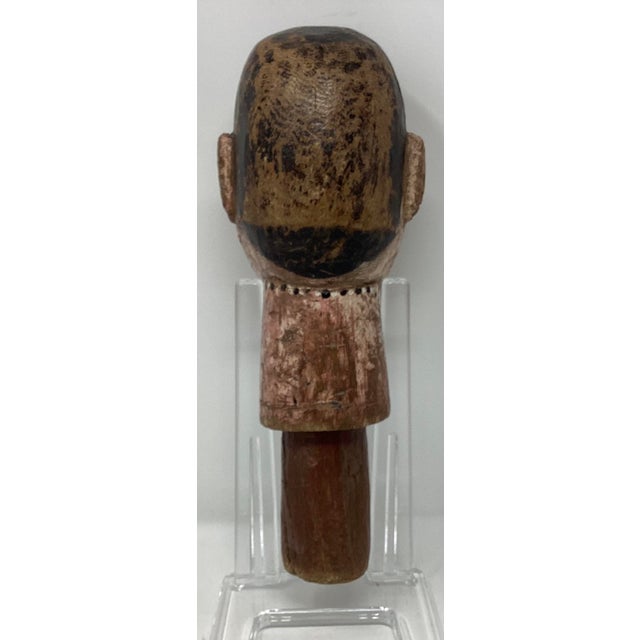 This carved and painted wooden sculpture features a stylized human face with prominent inlaid eyes, a red dot between the...
