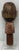 This carved and painted wooden sculpture features a stylized human face with prominent inlaid eyes, a red dot between the...
