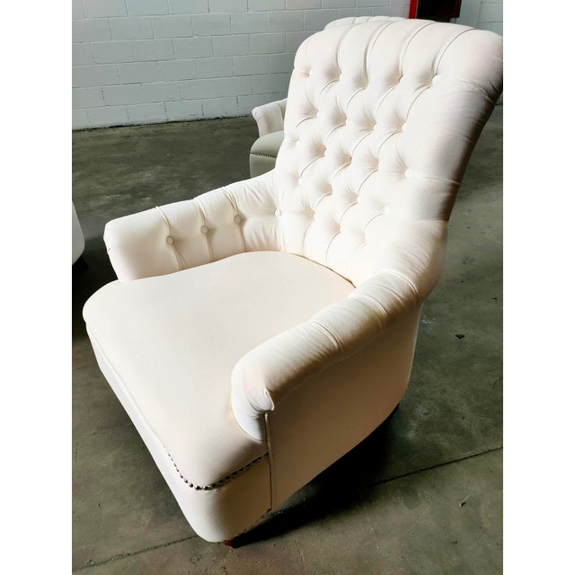 Armchairs with wooden frames and white fabric upholstery, button tufting on the back and arms, and studs on the bottom....