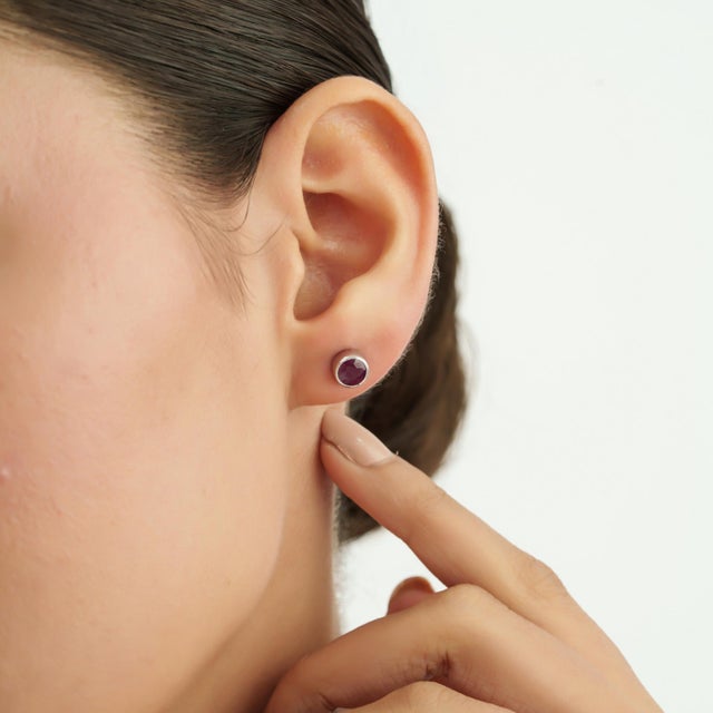 The Artisan Round Ruby Anniversary Stud Earrings feature vibrant round ruby gemstones with rich red hues and a faceted...